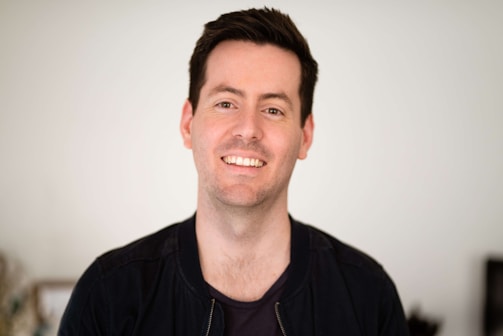 A person with short dark hair is smiling, wearing a dark jacket against a plain light background.