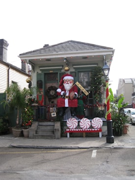 A house is decorated for the Christmas season with a large Santa Claus figure sitting on the porch. The surrounding area is adorned with festive decorations, including a Christmas tree, colorful lights, and giant candy decorations resembling peppermint swirls. A 'Santa Claus Day' banner is displayed on the porch.