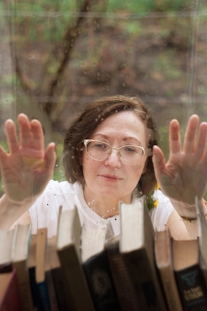 A person wearing glasses is looking through a rain-speckled window with hands gently pressed against it. The interior features a row of books in the foreground, creating a sense of longing or contemplation.