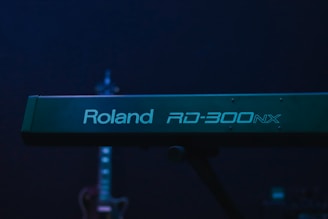 A Roland RD-300NX keyboard is prominent in the foreground, with its logo clearly visible. In the background, a guitar is slightly out of focus, adding depth to the image. The overall lighting is dim with a blue-green hue.
