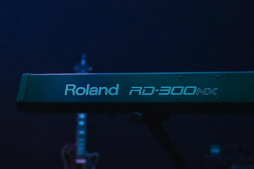 A Roland RD-300NX keyboard is prominent in the foreground, with its logo clearly visible. In the background, a guitar is slightly out of focus, adding depth to the image. The overall lighting is dim with a blue-green hue.
