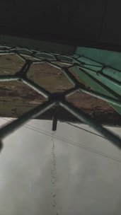 A fence with a hexagonal pattern viewed from below, with rainwater dripping from a pipe that protrudes above. The sky is overcast and there are electrical cables visible in the background.