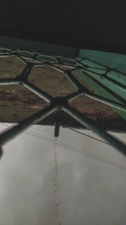 A fence with a hexagonal pattern viewed from below, with rainwater dripping from a pipe that protrudes above. The sky is overcast and there are electrical cables visible in the background.