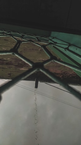A fence with a hexagonal pattern viewed from below, with rainwater dripping from a pipe that protrudes above. The sky is overcast and there are electrical cables visible in the background.