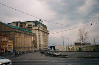 An urban scene featuring large buildings with intricate architecture, including a prominent multi-story structure with arched windows and a sloped roof. The street is curved with minimal traffic, showing a few vehicles. There is a mural wall on the right with various artworks, adjacent to a barren tree. The sky is overcast, adding a somber feel to the setting.