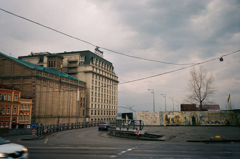 An urban scene featuring large buildings with intricate architecture, including a prominent multi-story structure with arched windows and a sloped roof. The street is curved with minimal traffic, showing a few vehicles. There is a mural wall on the right with various artworks, adjacent to a barren tree. The sky is overcast, adding a somber feel to the setting.