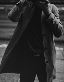 A monochrome photograph capturing a person wearing a long coat, with the coat partially open. The person is also wearing gloves and their face is not visible, adding an element of anonymity.
