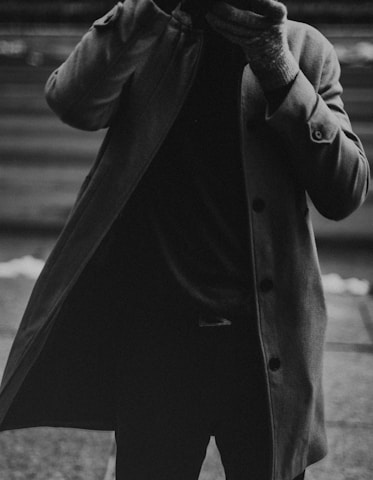 A monochrome photograph capturing a person wearing a long coat, with the coat partially open. The person is also wearing gloves and their face is not visible, adding an element of anonymity.