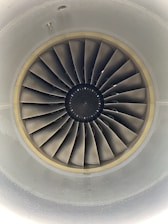 A close-up view of an aircraft jet engine turbine, featuring multiple metal blades arranged in a circular pattern. The design is symmetrical and technical, with shades of grey dominating the components. The intricate details of the blades and the central hub are clearly visible.