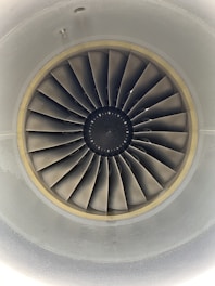 A close-up view of an aircraft jet engine turbine, featuring multiple metal blades arranged in a circular pattern. The design is symmetrical and technical, with shades of grey dominating the components. The intricate details of the blades and the central hub are clearly visible.