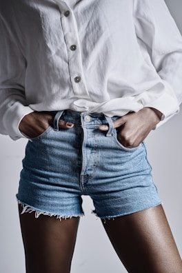 A person wearing a white button-up shirt and blue denim shorts with frayed edges. The hands are placed in the pockets, and the shirt is slightly oversized.