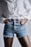A person wearing a white button-up shirt and blue denim shorts with frayed edges. The hands are placed in the pockets, and the shirt is slightly oversized.