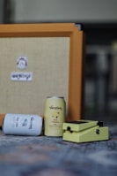 Two cans labeled 'Vacation' are placed in front of an orange amplifier. One can is lying down, displaying the label 'Day Spa,' while the other stands upright with the label 'Hot Tropics.' A yellow guitar effects pedal is positioned beside the cans.