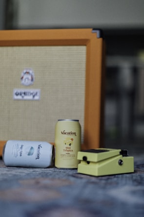 Two cans labeled 'Vacation' are placed in front of an orange amplifier. One can is lying down, displaying the label 'Day Spa,' while the other stands upright with the label 'Hot Tropics.' A yellow guitar effects pedal is positioned beside the cans.