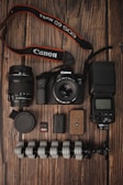 A collection of photography equipment is neatly arranged on a wooden surface. Items include a Canon DSLR camera with a lens attached, an additional lens, a camera strap, memory cards, battery, a flexible tripod, a camera flash unit, and a lens cap.