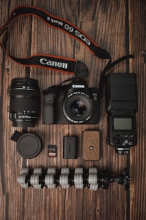 A collection of photography equipment is neatly arranged on a wooden surface. Items include a Canon DSLR camera with a lens attached, an additional lens, a camera strap, memory cards, battery, a flexible tripod, a camera flash unit, and a lens cap.