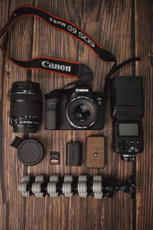 A collection of photography equipment is neatly arranged on a wooden surface. Items include a Canon DSLR camera with a lens attached, an additional lens, a camera strap, memory cards, battery, a flexible tripod, a camera flash unit, and a lens cap.