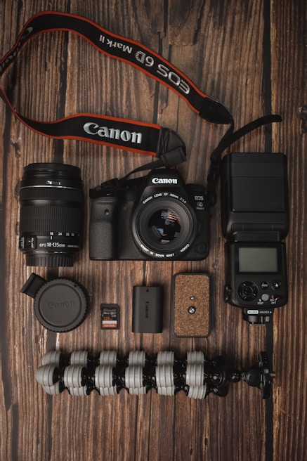 A collection of photography equipment is neatly arranged on a wooden surface. Items include a Canon DSLR camera with a lens attached, an additional lens, a camera strap, memory cards, battery, a flexible tripod, a camera flash unit, and a lens cap.