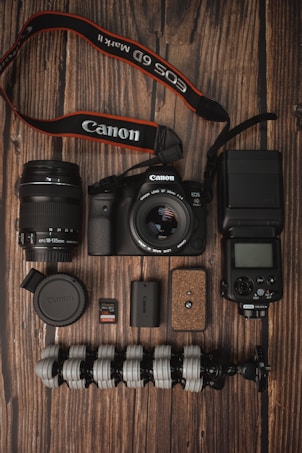 A collection of photography equipment is neatly arranged on a wooden surface. Items include a Canon DSLR camera with a lens attached, an additional lens, a camera strap, memory cards, battery, a flexible tripod, a camera flash unit, and a lens cap.