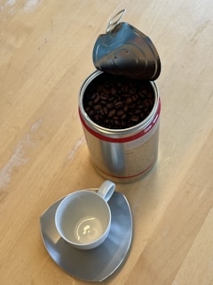 A metal container filled with dark roasted coffee beans is open, placed on a light wooden surface. Beside it, a white coffee cup sits atop an unconventional triangular saucer, both with a glossy finish.