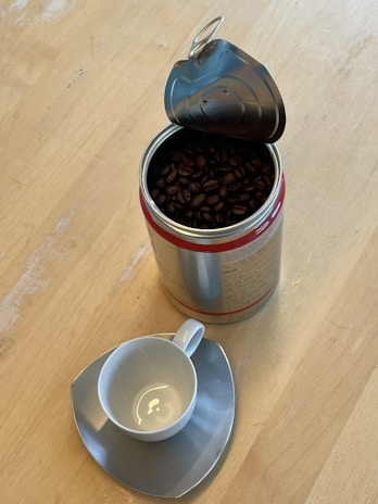 A metal container filled with dark roasted coffee beans is open, placed on a light wooden surface. Beside it, a white coffee cup sits atop an unconventional triangular saucer, both with a glossy finish.
