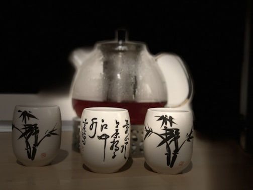 A glass teapot filled with tea is placed in the background, with three ceramic cups decorated with black Asian-style calligraphy and bamboo designs in the foreground. The scene is set on a wooden surface against a dark background, which emphasizes the brightness and details of the teapot and cups.