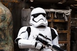 A person wearing a white futuristic armor suit with black accents, holding a large blaster weapon. The background includes a rustic, industrial setting with wooden and metallic elements.