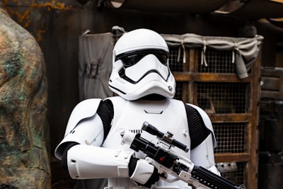 A person wearing a white futuristic armor suit with black accents, holding a large blaster weapon. The background includes a rustic, industrial setting with wooden and metallic elements.