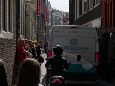 A narrow city street in a European urban area with pedestrians standing on the sidewalk next to a parked van. The van is white with multiple brand logos and the word 'TURQUOISE' written on it. A person on a scooter or motorcycle is riding closely behind the van. The buildings on either side are tall and closely packed, some with signs in Dutch.