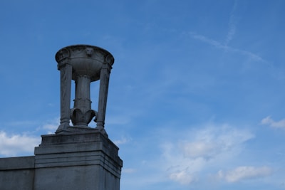 A stone urn with ornate carvings sits atop a concrete structure against a clear blue sky. The urn is detailed with vertical grooves and decorative patterns.