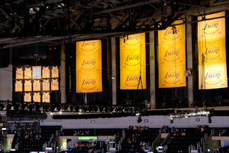 A sports arena interior featuring several large yellow banners hanging from the ceiling, displaying 'Los Angeles Lakers' with championship years. Below, retired jersey numbers are visible, commemorating past players. In the background, sections of seating contain scattered groups of people.