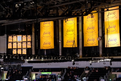 A sports arena interior featuring several large yellow banners hanging from the ceiling, displaying 'Los Angeles Lakers' with championship years. Below, retired jersey numbers are visible, commemorating past players. In the background, sections of seating contain scattered groups of people.