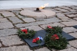 An eternal flame burns on a circular stone platform surrounded by a tiled stone ground. Several wreaths made of green foliage and adorned with red ribbons are placed on dark rectangular slabs near the flame.