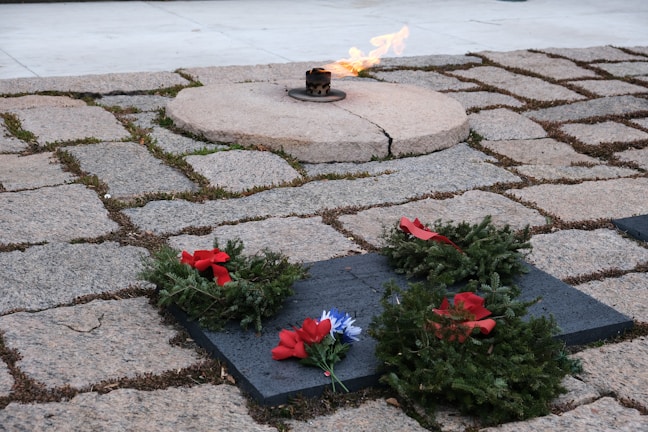 An eternal flame burns on a circular stone platform surrounded by a tiled stone ground. Several wreaths made of green foliage and adorned with red ribbons are placed on dark rectangular slabs near the flame.