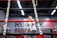 A person is training inside a boxing ring, with 'Everlast' branding visible on the ropes. The focus seems to be on the foreground ropes, while the person appears slightly blurred in the background. The setting includes a gym-like environment with dim lighting and industrial design elements.