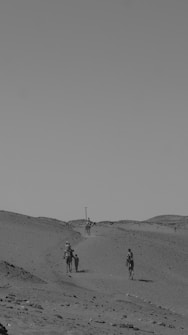 A monochrome scene featuring a desert landscape with gentle rolling sand dunes. Several people ride camels, traveling along a path that weaves through the dunes. The sky is clear, adding to the vastness and openness of the environment.