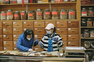 Two people wearing masks and gloves work behind a counter in an herbal medicine shop, surrounded by numerous wooden drawers and jars filled with various herbs and ingredients.