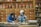 Two people wearing masks and gloves work behind a counter in an herbal medicine shop, surrounded by numerous wooden drawers and jars filled with various herbs and ingredients.