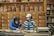 Two people wearing masks and gloves work behind a counter in an herbal medicine shop, surrounded by numerous wooden drawers and jars filled with various herbs and ingredients.
