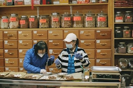 Two people wearing masks and gloves work behind a counter in an herbal medicine shop, surrounded by numerous wooden drawers and jars filled with various herbs and ingredients.
