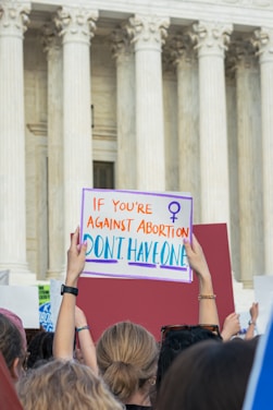 A group of people are gathered outside a large, white stone building with Greek-style columns. One person is visibly holding a sign that reads 'If you're against abortion, don't have one' with a female gender symbol. The crowd appears to be part of a protest or demonstration.