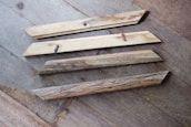 Four wooden planks are lying on a wooden floor. The planks have a natural, rustic finish, and two of them have screws partially embedded. The floor has a variegated pattern with a mix of light and dark brown tones.