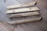 Four wooden planks are lying on a wooden floor. The planks have a natural, rustic finish, and two of them have screws partially embedded. The floor has a variegated pattern with a mix of light and dark brown tones.