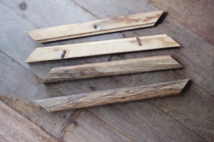 Four wooden planks are lying on a wooden floor. The planks have a natural, rustic finish, and two of them have screws partially embedded. The floor has a variegated pattern with a mix of light and dark brown tones.