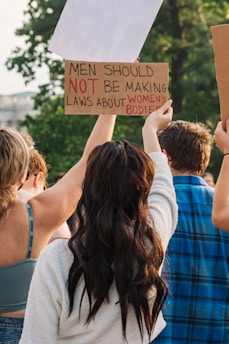 A group of people participates in a protest, holding signs in the air. One sign reads 'Men should not be making laws about women's bodies' with certain words emphasized in red. The image is set outdoors with green foliage in the background.