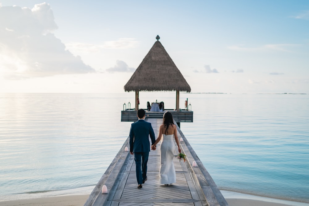 a man and woman walking down a dock towards a hut on the water