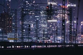 A nighttime urban scene featuring the infrastructure of train tracks and overhead wires against the backdrop of a city skyline filled with illuminated skyscrapers. Traffic lights and signs are visible along the railway lines.
