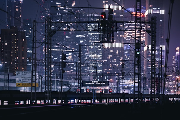 A nighttime urban scene featuring the infrastructure of train tracks and overhead wires against the backdrop of a city skyline filled with illuminated skyscrapers. Traffic lights and signs are visible along the railway lines.