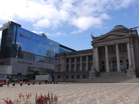 Glass-fronted modern building with Microsoft and Nordstrom signage adjacent to an older classical-style building with columns and a dome. A few people and planters are visible on the wide paved area in the foreground under a partly cloudy sky.