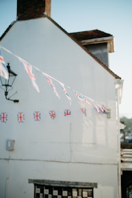 A white building with a brick chimney features a string of Union Jack flags hanging across its facade. A traditional black metal streetlamp is affixed on the side of the building near the flags. The lower part of the building shows a window with a black and white checker pattern. The sky is clear and blue, indicating daylight.